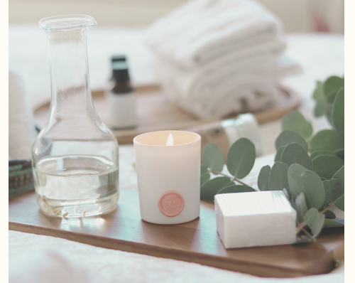 Spa setup with a tray of relaxation items including a lit candle, eucalyptus leaves, a bar of soap, a glass carafe of water, and rolled white towels.