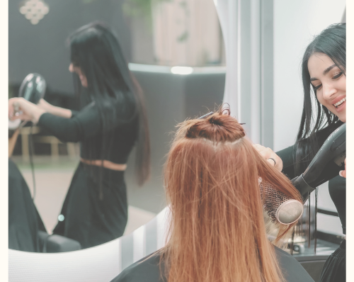 Hairdresser styling a client's red hair with a blow dryer in a salon, with a mirror reflecting the process and a modern salon setup in the background.