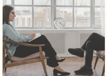 Therapy session with two women sitting in a cozy room, one taking notes and the other speaking, with a clock on the wall and large windows in the background.