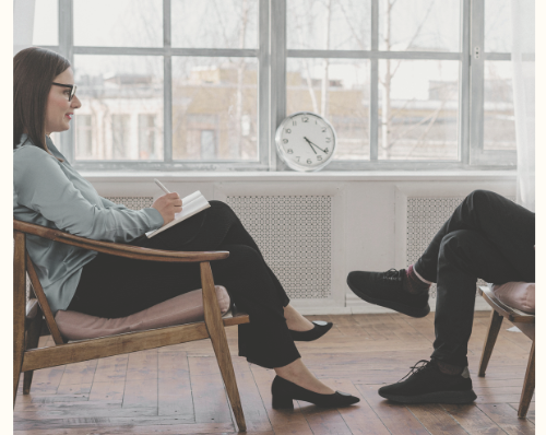 Therapy session with two women sitting in a cozy room, one taking notes and the other speaking, with a clock on the wall and large windows in the background.
