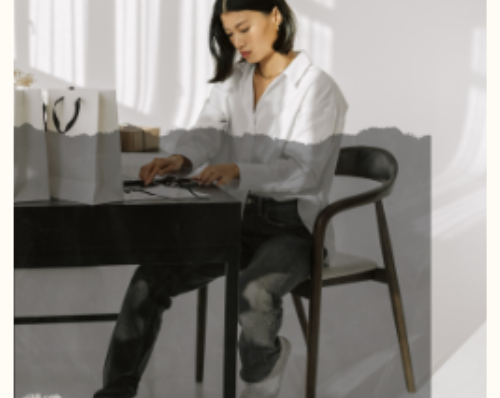 Woman working at a desk in a modern office, writing in a notebook with shopping bags nearby, in a bright and airy workspace.