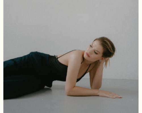 Woman in a black dress lying on the floor, resting her head on her hand, in a minimalist setting with a neutral background.