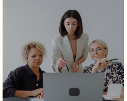 Three women in a business meeting, gathered around a laptop, discussing ideas in a professional setting with notebooks on the table.
