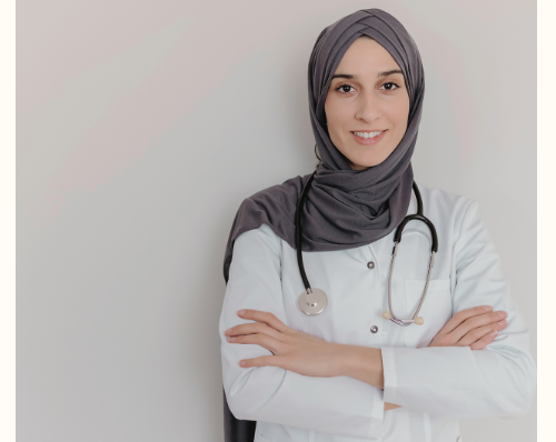 Female doctor wearing a hijab and white coat with a stethoscope around her neck, standing with arms crossed and smiling confidently.